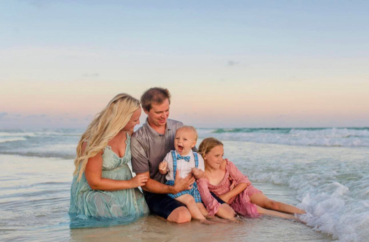 A young mom and dad sitting at the water's edge holding their small son and their daughter laying beside them