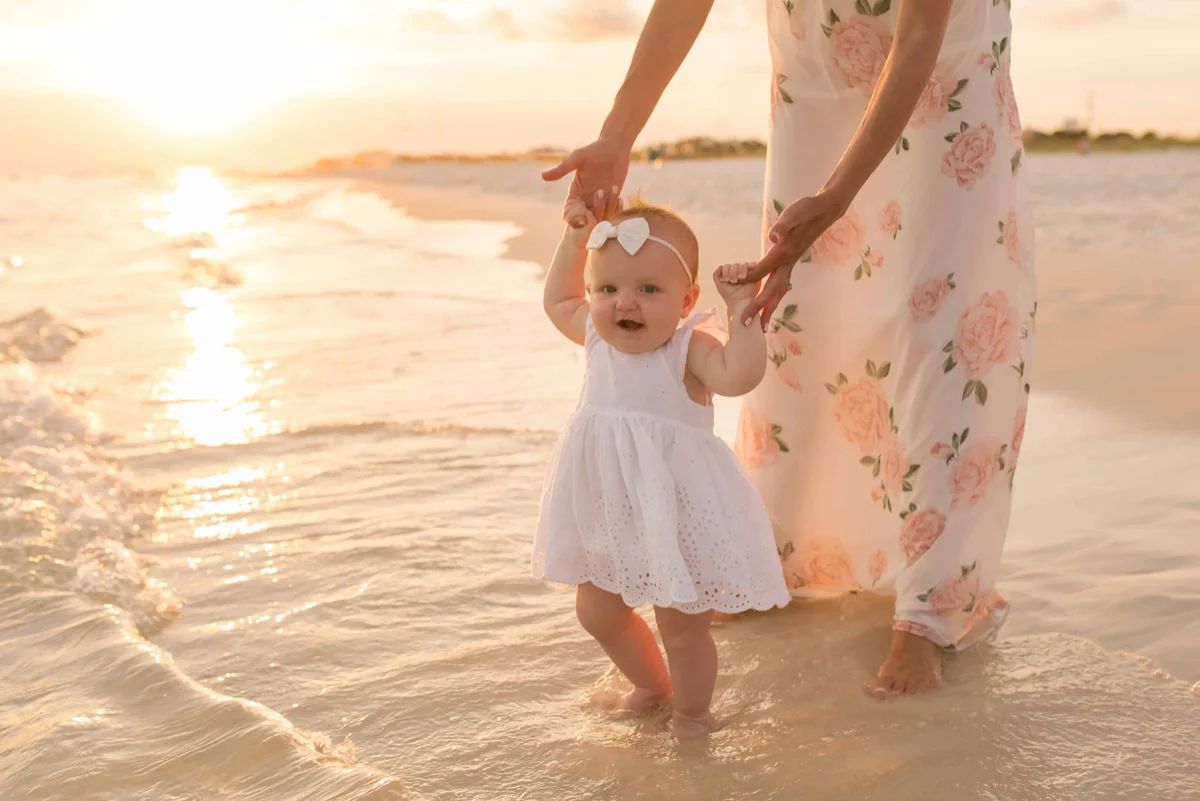 A baby in a white dress with a white bow, holding onto her mama's hands while standing in the water at the shoreline