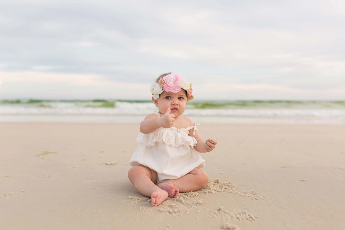Baby with white dress and floral headband, sitting on beach, with water in background.