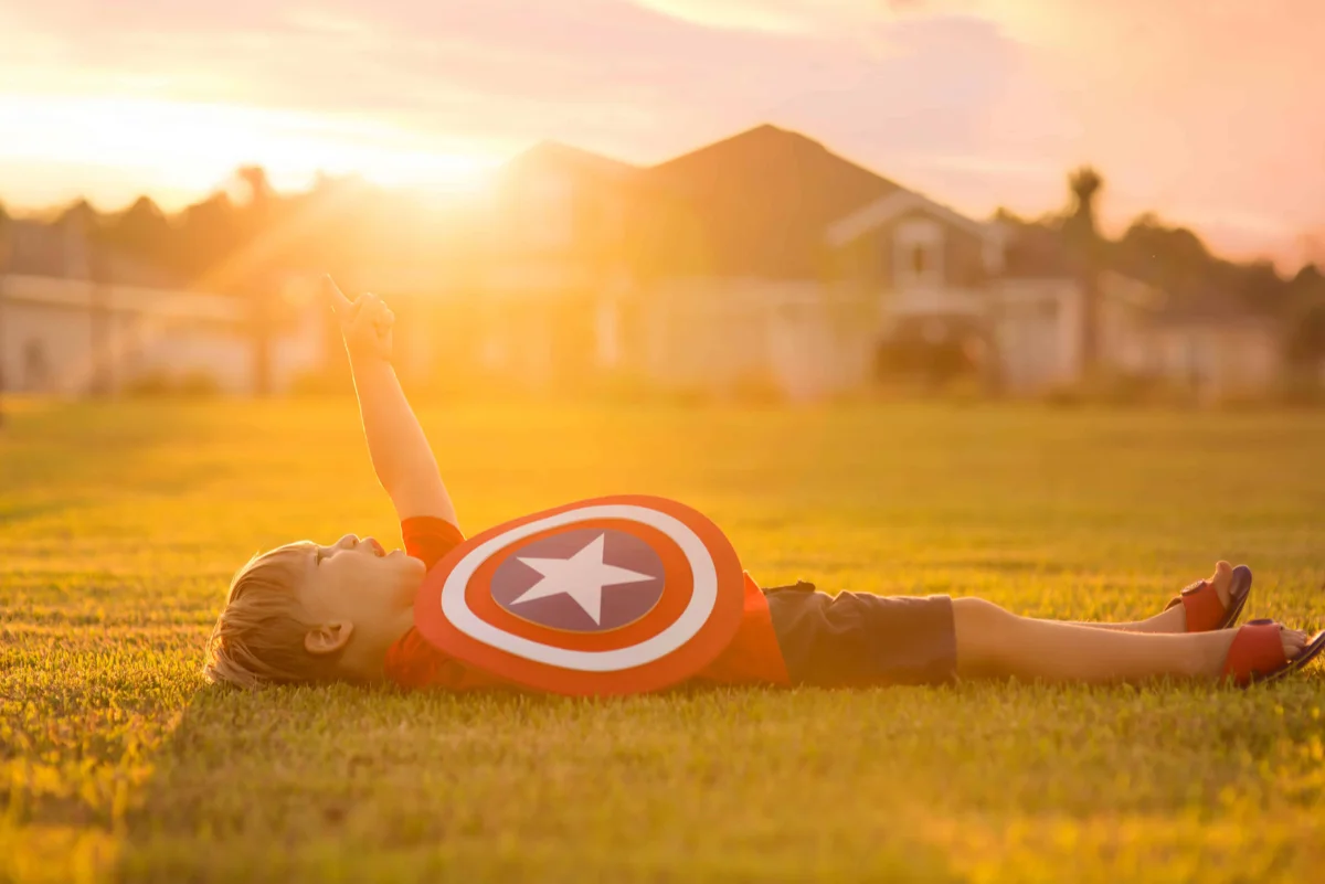A young boy laying in the grass, staring up at the sky, holding a Captain America shield,