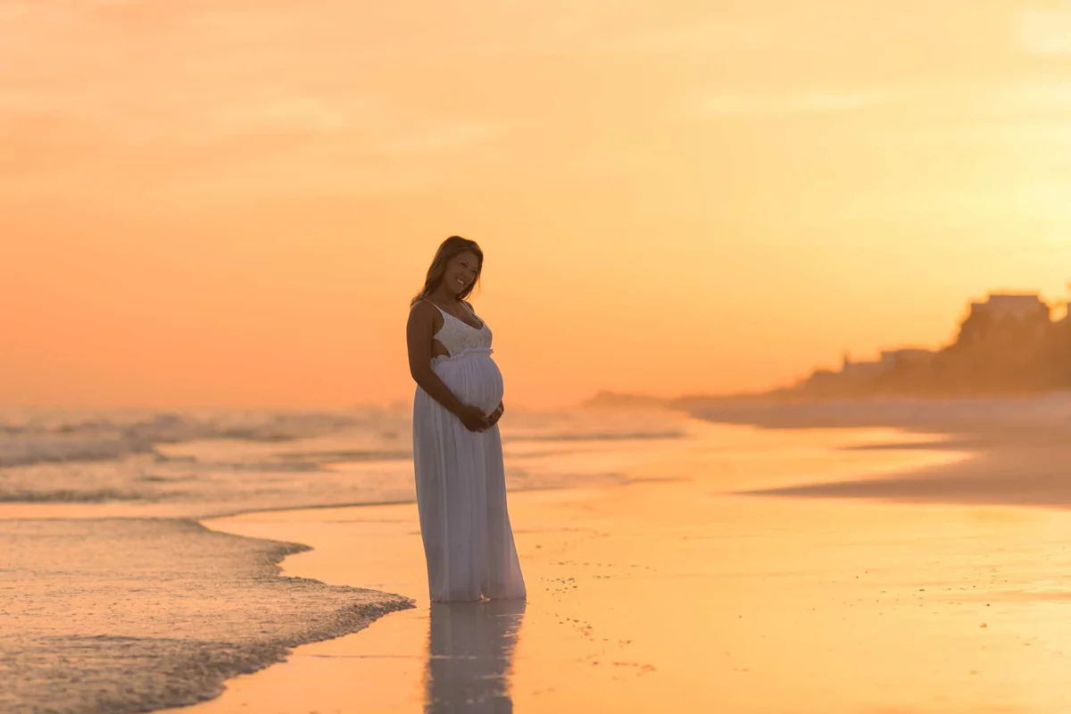 A mother to be in a long flowing white dress, in the sunset at the water's edge