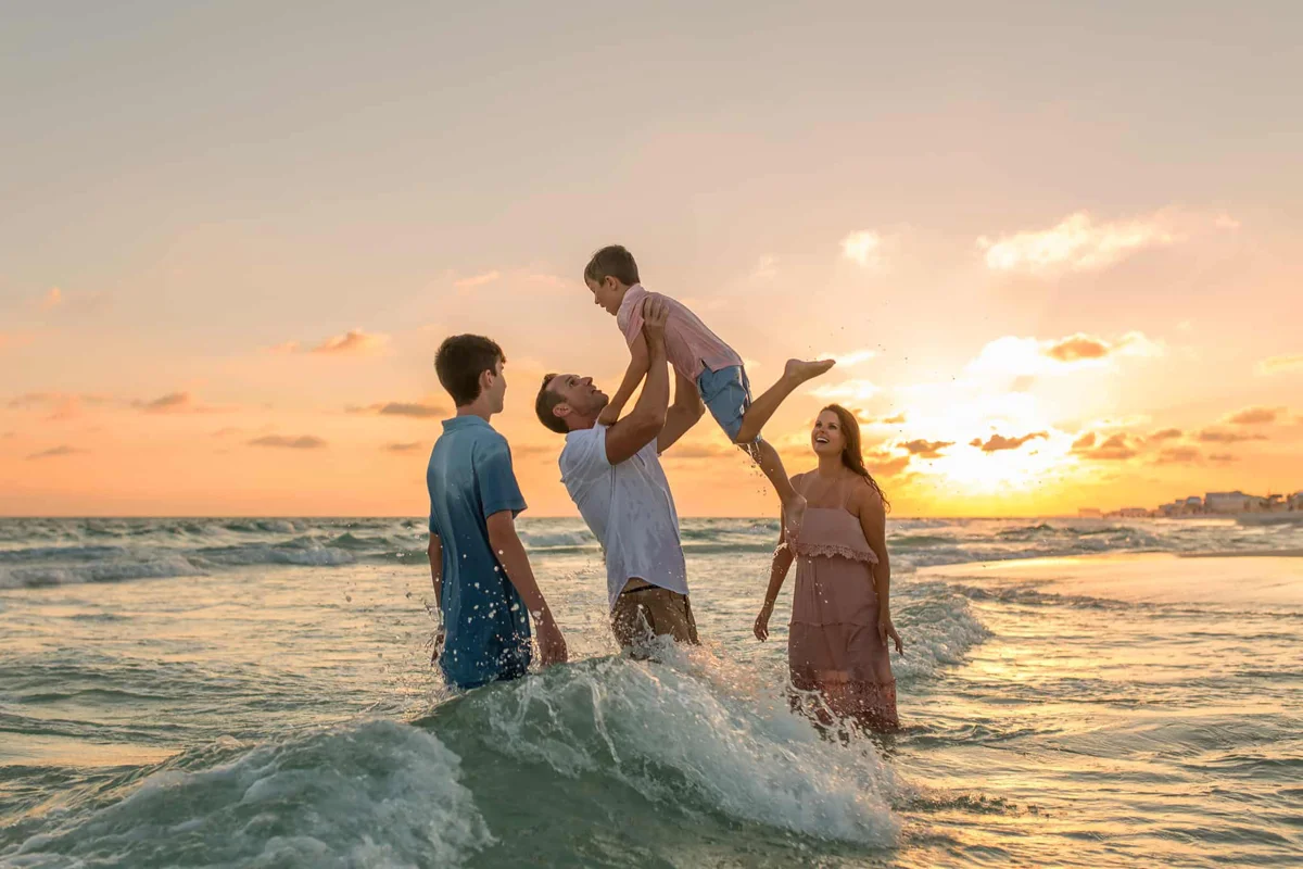 faq A mother and father playing in the ocean at sunset with their two sons.