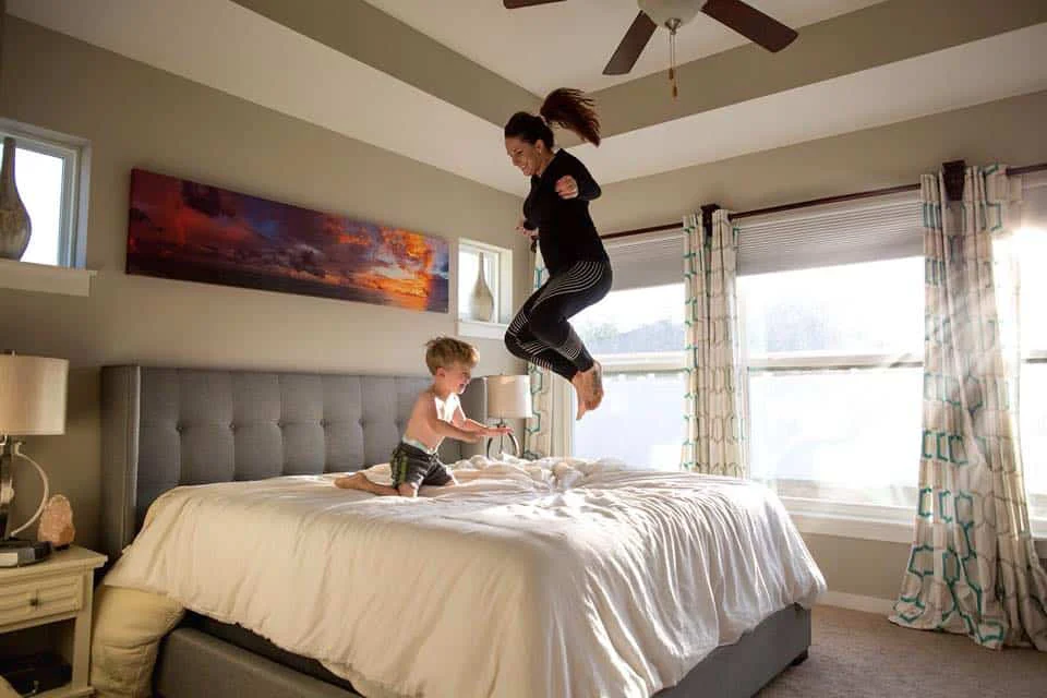 A mom jumpimg on a bed, while playing with her young son.