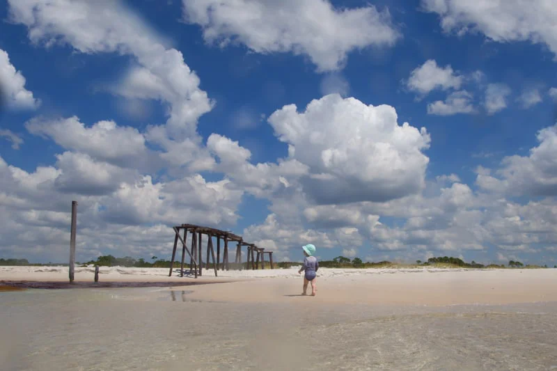 A small child in a sun hat walking on the beach's water edge. An old dock in the background and a sky full of fluffy clouds.