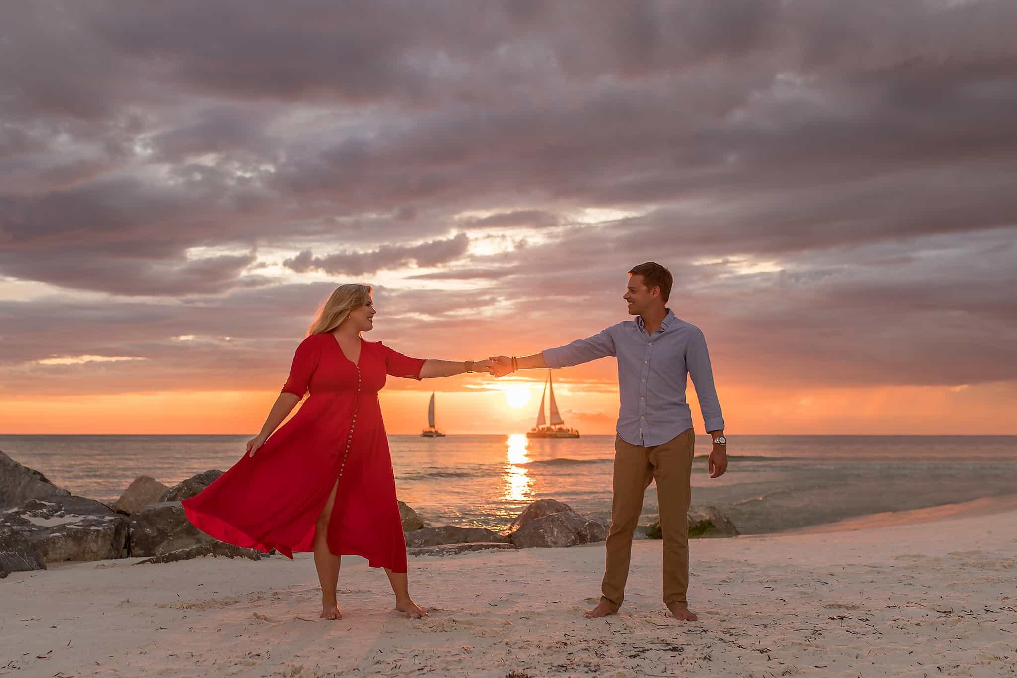A couple dancing together on the beach at sunset. The female is wearing a red midi dress, the male is wearing long pants and a long sleeved shirt
